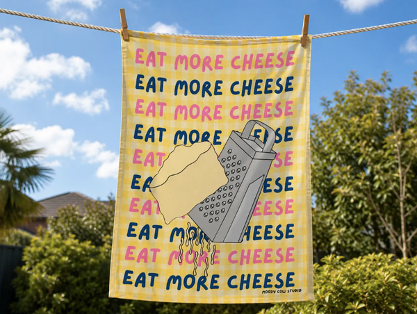 Towel with 'Eat More Cheese' text and a grater graphic hanging on a line outdoors.